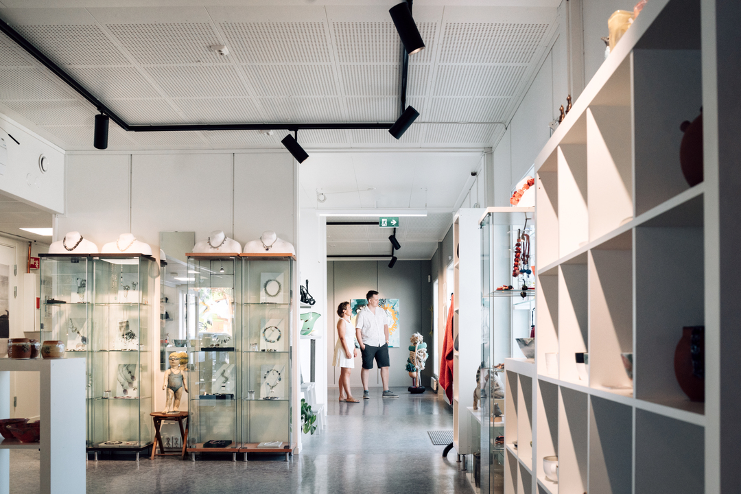 Ett ljust galleri med konstföremål. I mitten av rummet står två besökare och tittar på en väggmonterad konstbild. Lokalen har vita väggar, hyllor med keramikföremål, spotbelysning i taket och en öppen, modern utformning. I Slemmestad centrum i Norge.