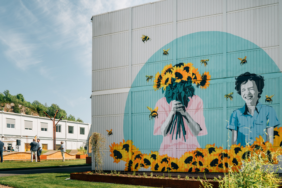 En stor färgglad muralmålning på en byggnadsfasad föreställer två personer omgivna av solrosor och flygande bin. Den ena håller en bukett solrosor framför ansiktet, medan den andra ler. Framför byggnaden finns gräs och planteringar, och till vänster syns människor som rör sig i ett öppet utomhusområde under en klar himmel. Slemmestad centrum i Norge.