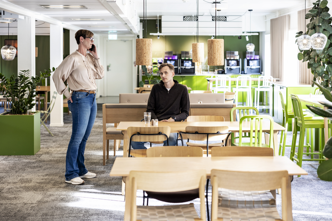 Ljus paus- och mötesyta i kontorsmoduler hos Polypeptide med träbord, gröna stolar, pendelbelysning och växter, där medarbetare vistas och samtalar i en modern och trivsam arbetsmiljö – kontorsmoduler från Expandia.