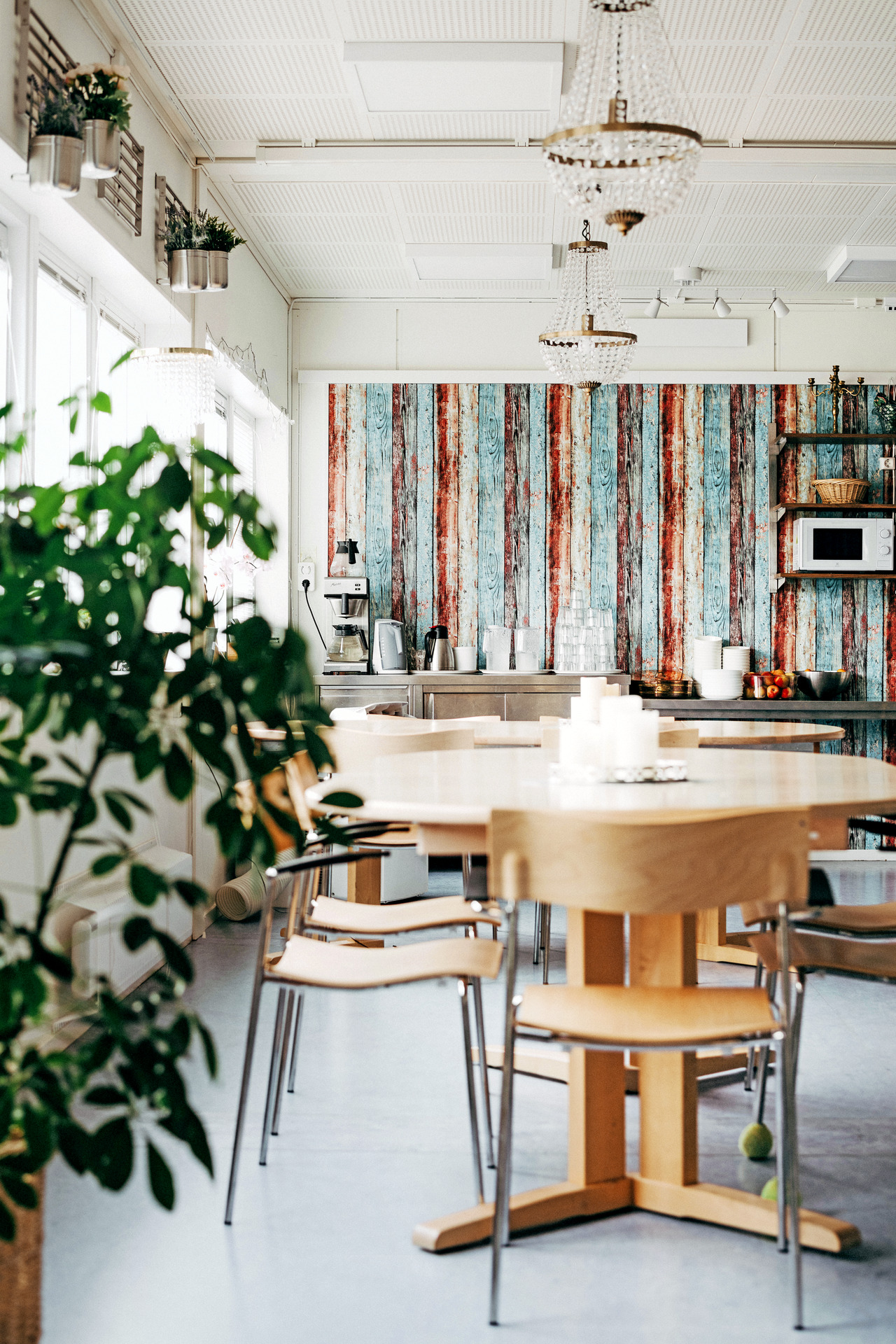 Matsal med runda träbord och stolar i ljusa toner. I bakgrunden syns en serveringsyta med kaffemaskin, glas och frukt. Rummet är ljust och ombonat med växter, dekorativa lampor och en färgglad fondvägg i trästruktur.
