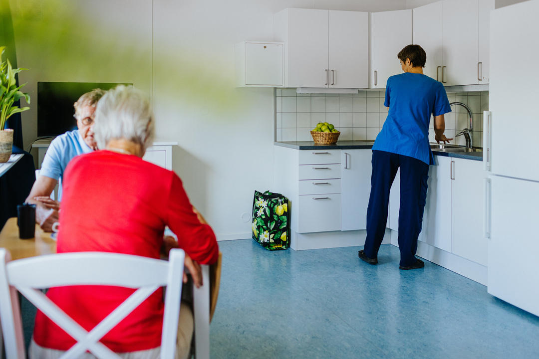 Inomhusmiljö vid Herrhagsgården vård- och omsorgsboende i Falun där två äldre personer sitter vid ett bord medan omsorgspersonal arbetar vid köksbänken. Rummet har ljusa ytor och ett funktionellt kök som ingår i boendets modullösning.