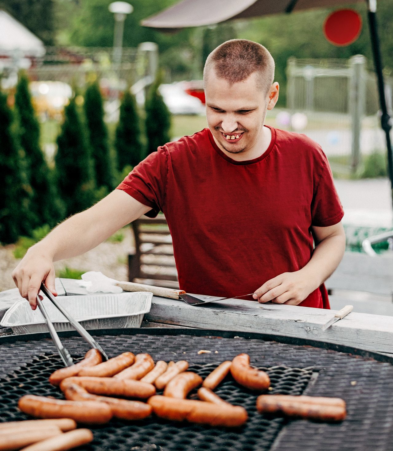 En person står utomhus och grillar korv, vänder korvarna med en tång och ler.